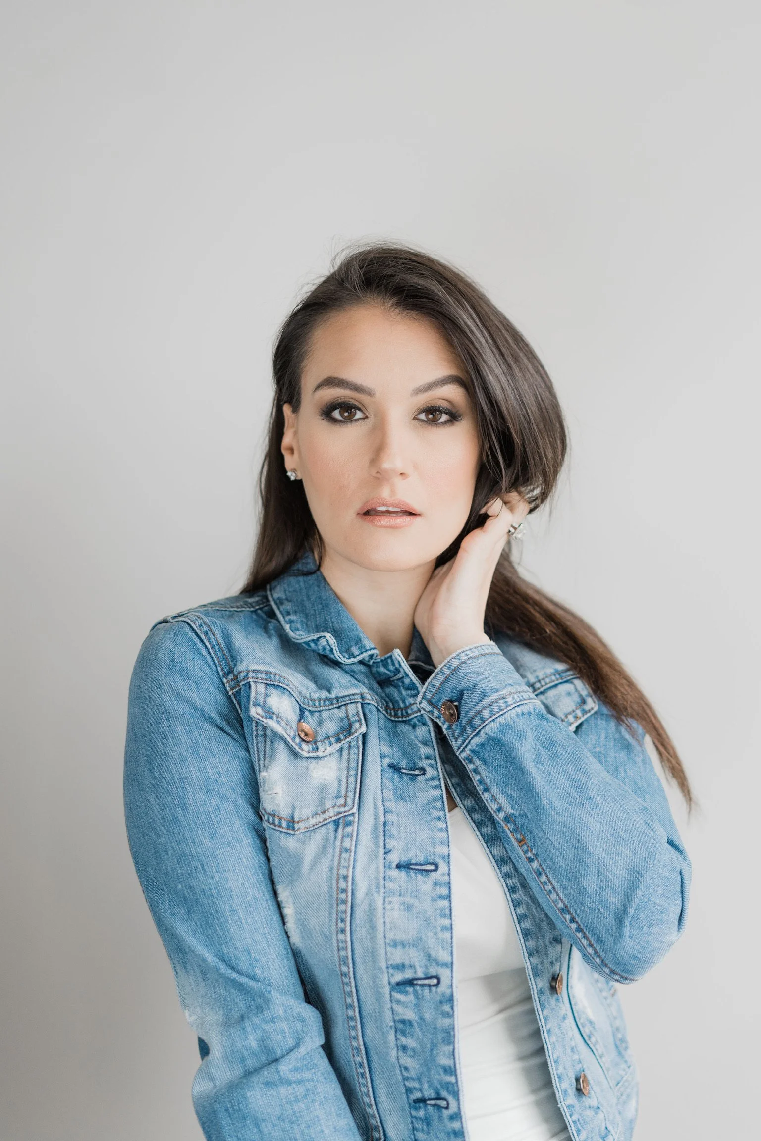 A woman with dark brown hair wearing a denim jacket and white top, posing against a plain light grey background.