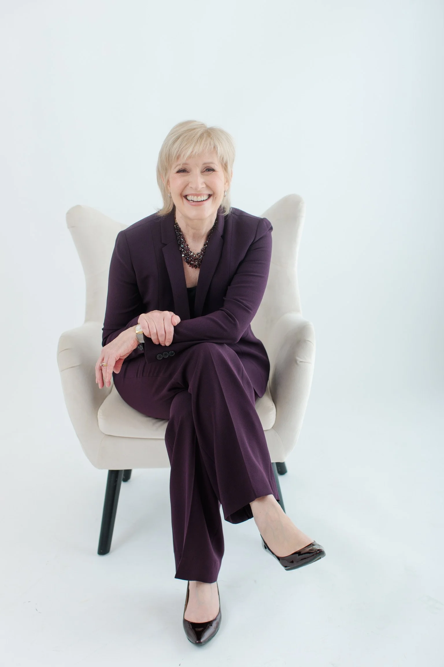 A woman with short blond hair, sitting on a cream armchair, smiling, wearing a dark purple suit and black high heels.