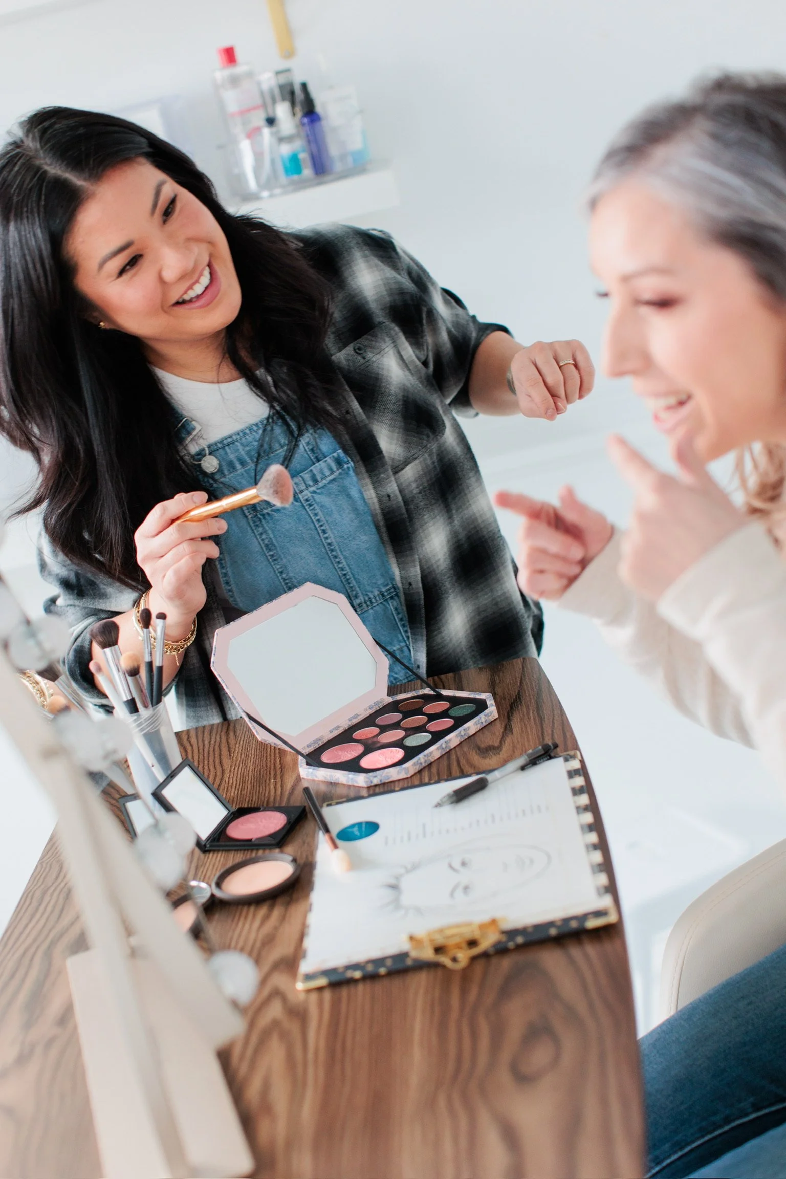 A makeup artist cheerfully applying makeup to a woman sitting at a vanity table with various makeup products, brushes, and a hand mirror.