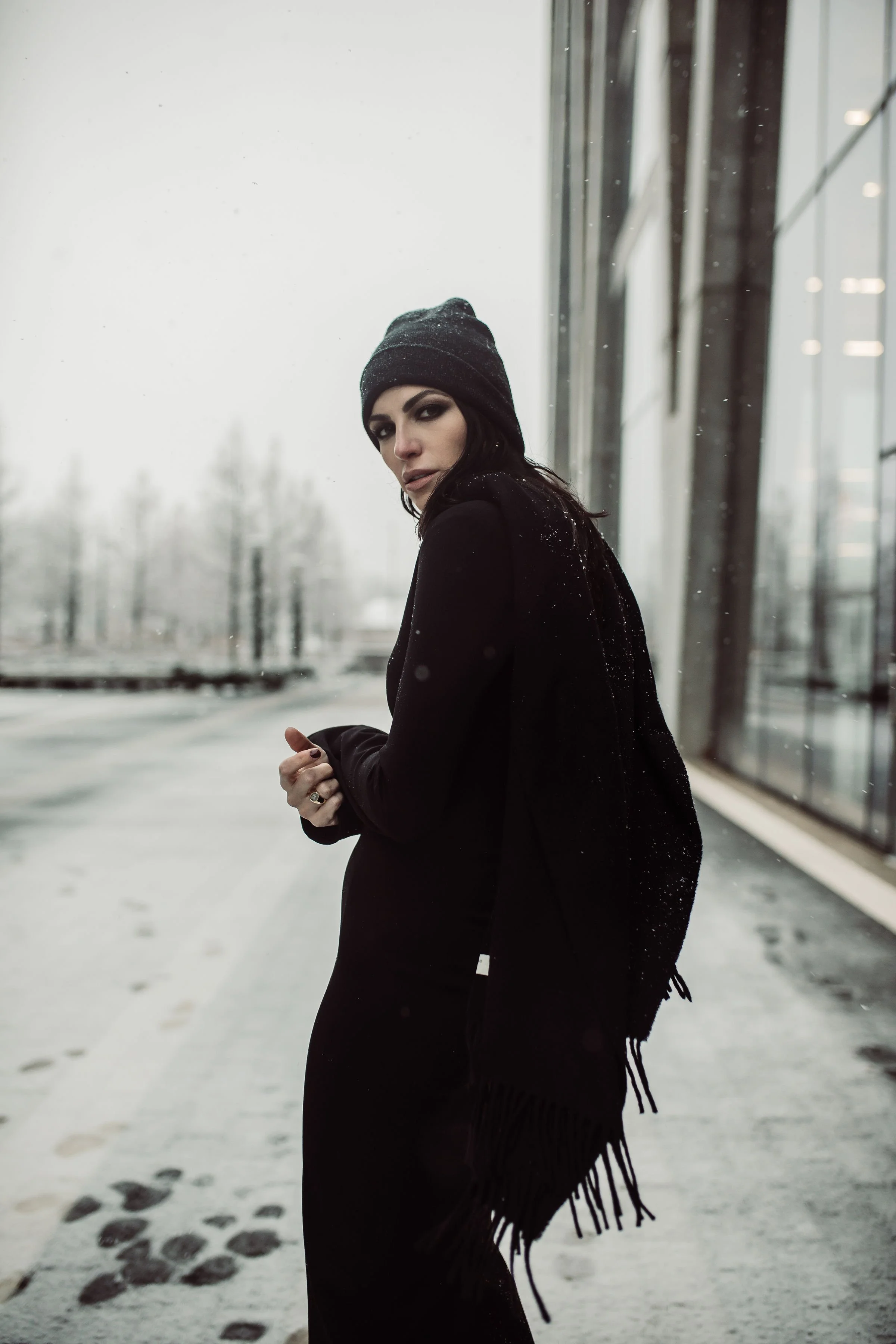 A woman wearing black clothing and a beanie hat standing outside in snowy weather beside a modern glass building.