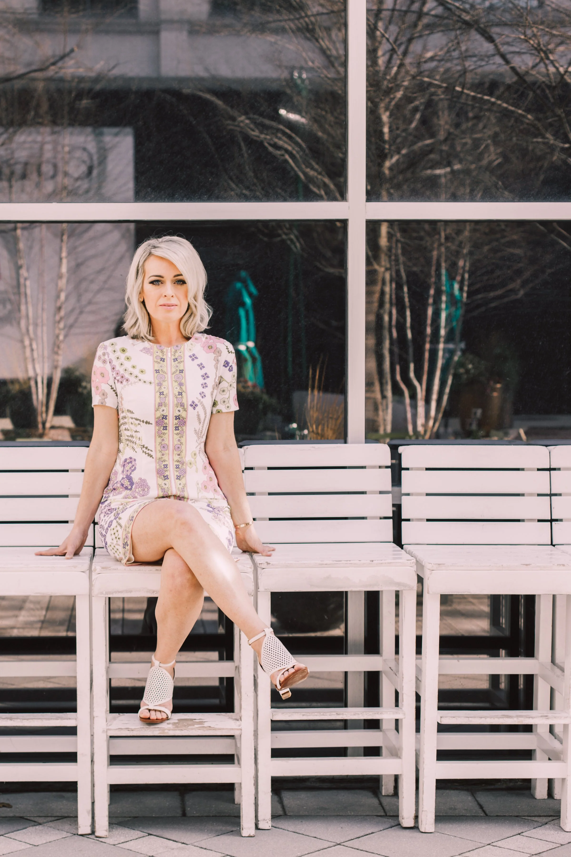 A woman with blonde hair sitting on a white bench outside, in front of a large window, wearing a floral dress and white heels.