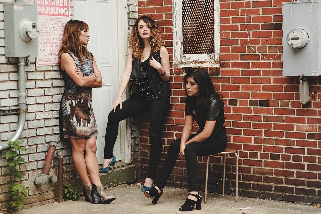 Three young women with dark makeup and fashionable clothing in an alleyway with brick walls. One woman stands against the wall with arms crossed, another leans against a door with one foot on the ground, and the third sits on a stool looking downward