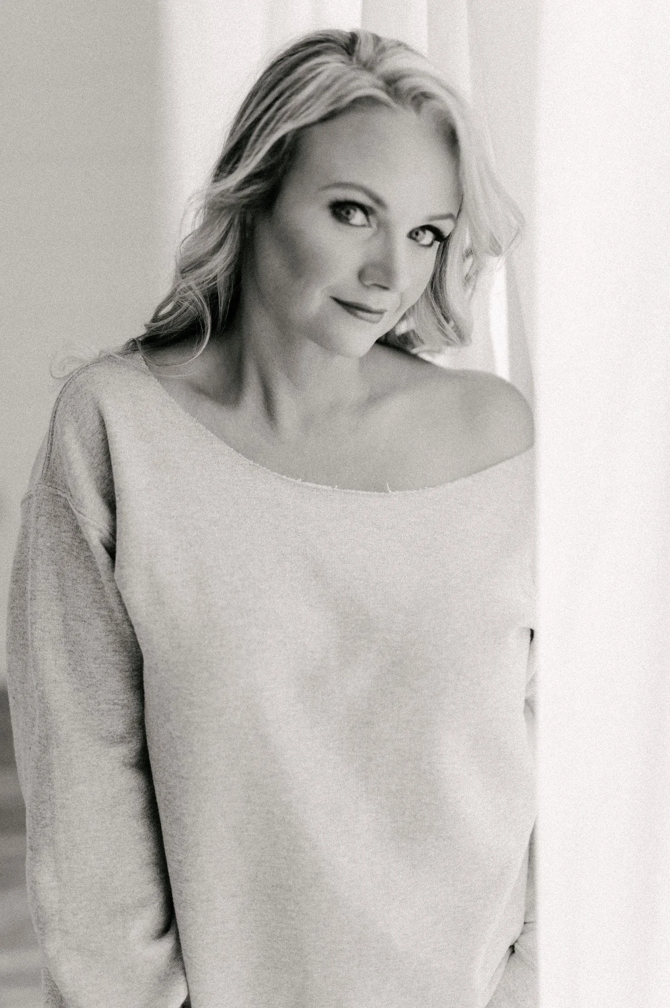 Black and white photo of a woman with shoulder-length hair, wearing an off-the-shoulder sweater, looking at the camera with a slight smile.