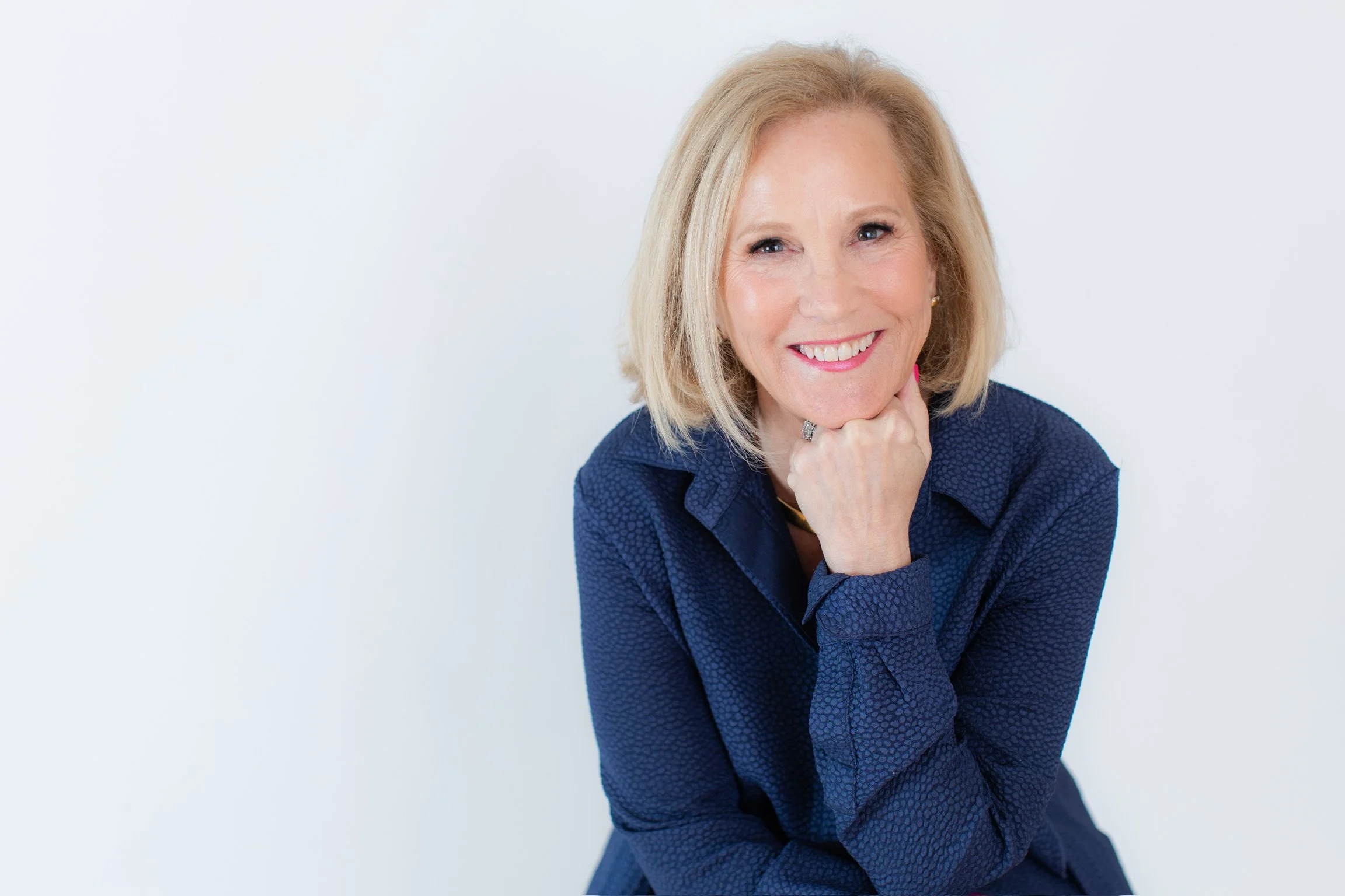 A smiling middle-aged woman with blonde hair, wearing a dark blue textured blouse, resting her chin on her hand against a plain white background.