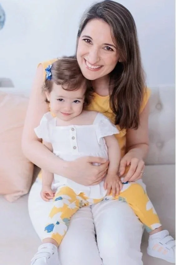 A woman with long brown hair and a yellow top smiling while sitting on a sofa, holding a small girl with brown hair in a white shirt with buttons and yellow pants with a floral pattern, both looking at the camera.