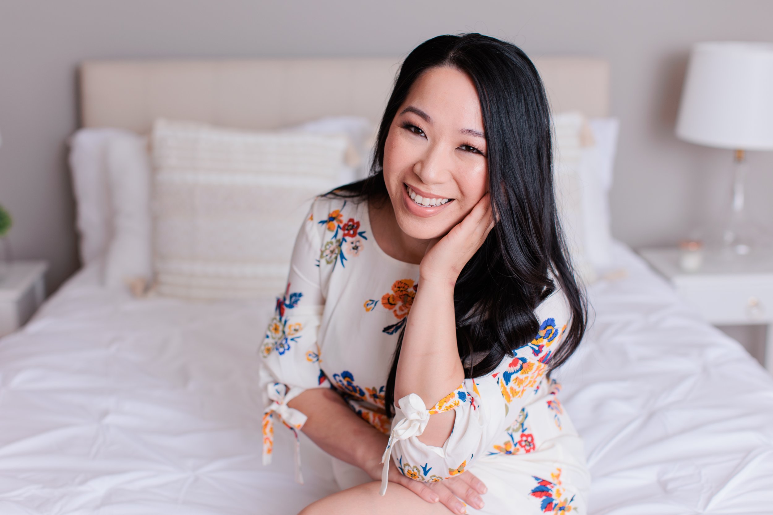 A smiling woman with long black hair, wearing a white floral dress, sitting on a bed in a bedroom with white bedding and a nightstand with a lamp.