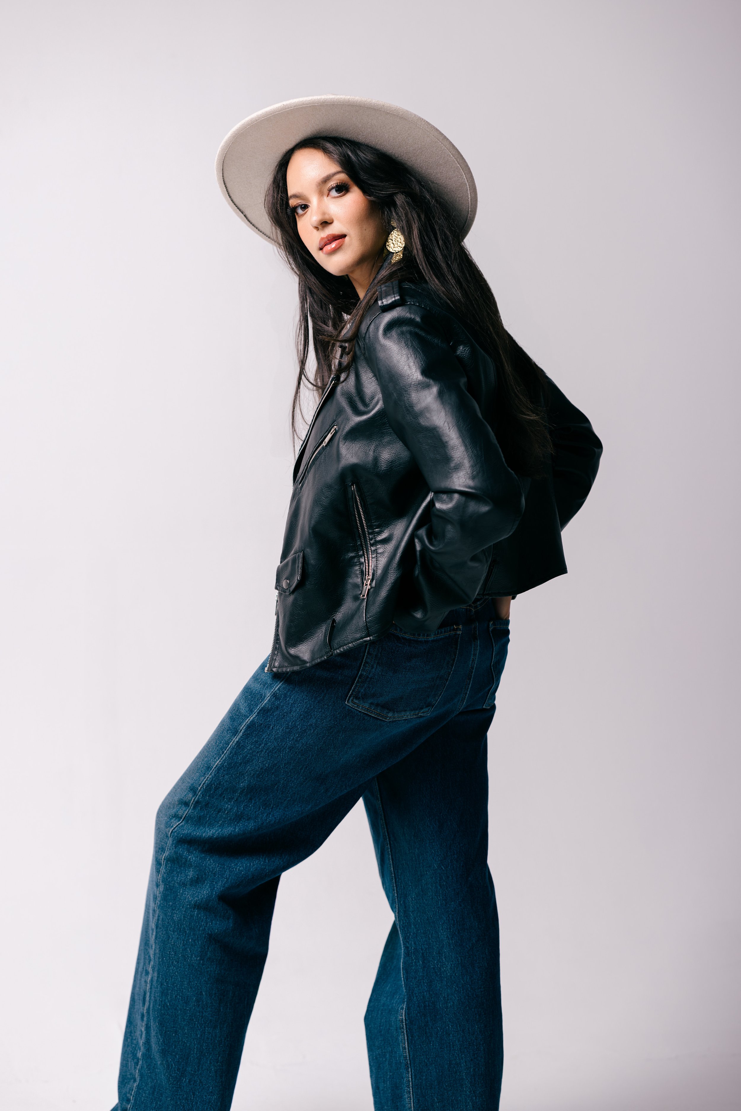 Woman wearing a black leather jacket, blue jeans, beige wide-brim hat, and gold earrings, standing against a light gray background.