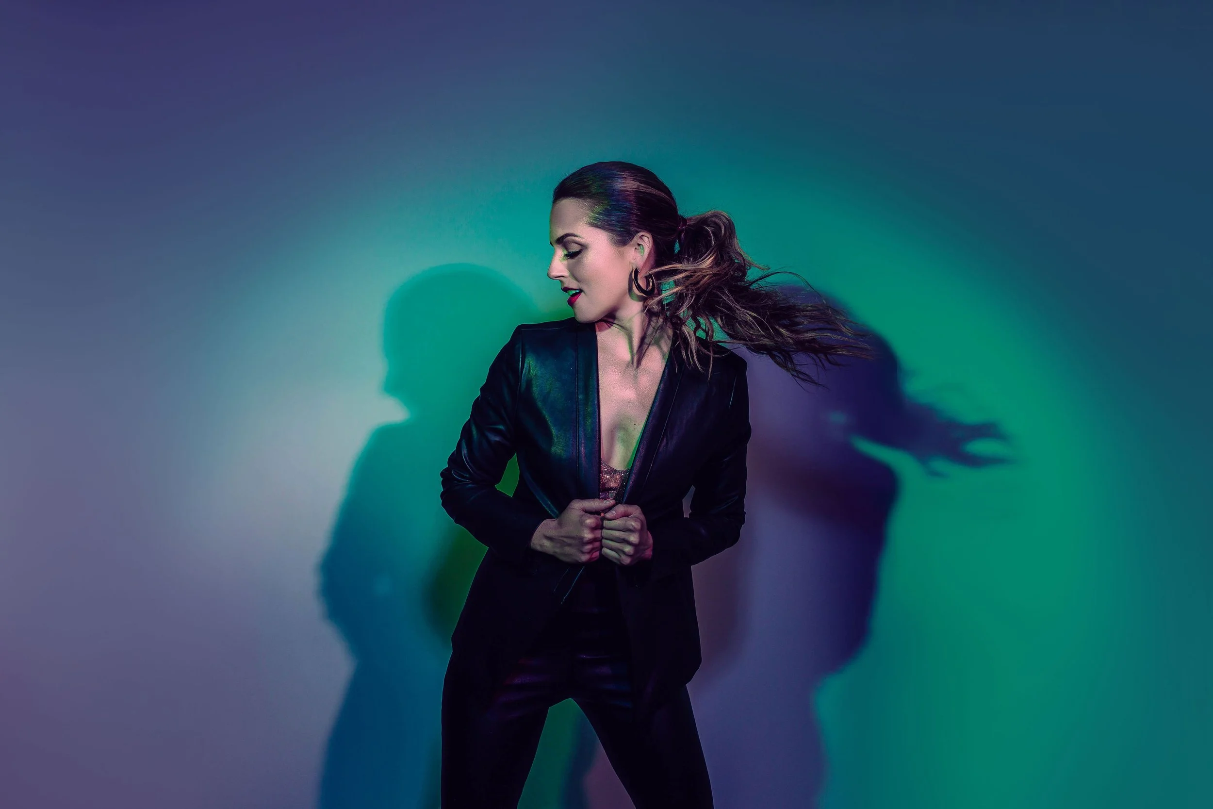 A woman with long hair in a ponytail, wearing a black leather jacket, stands against a wall with colorful lighting casting shadows.