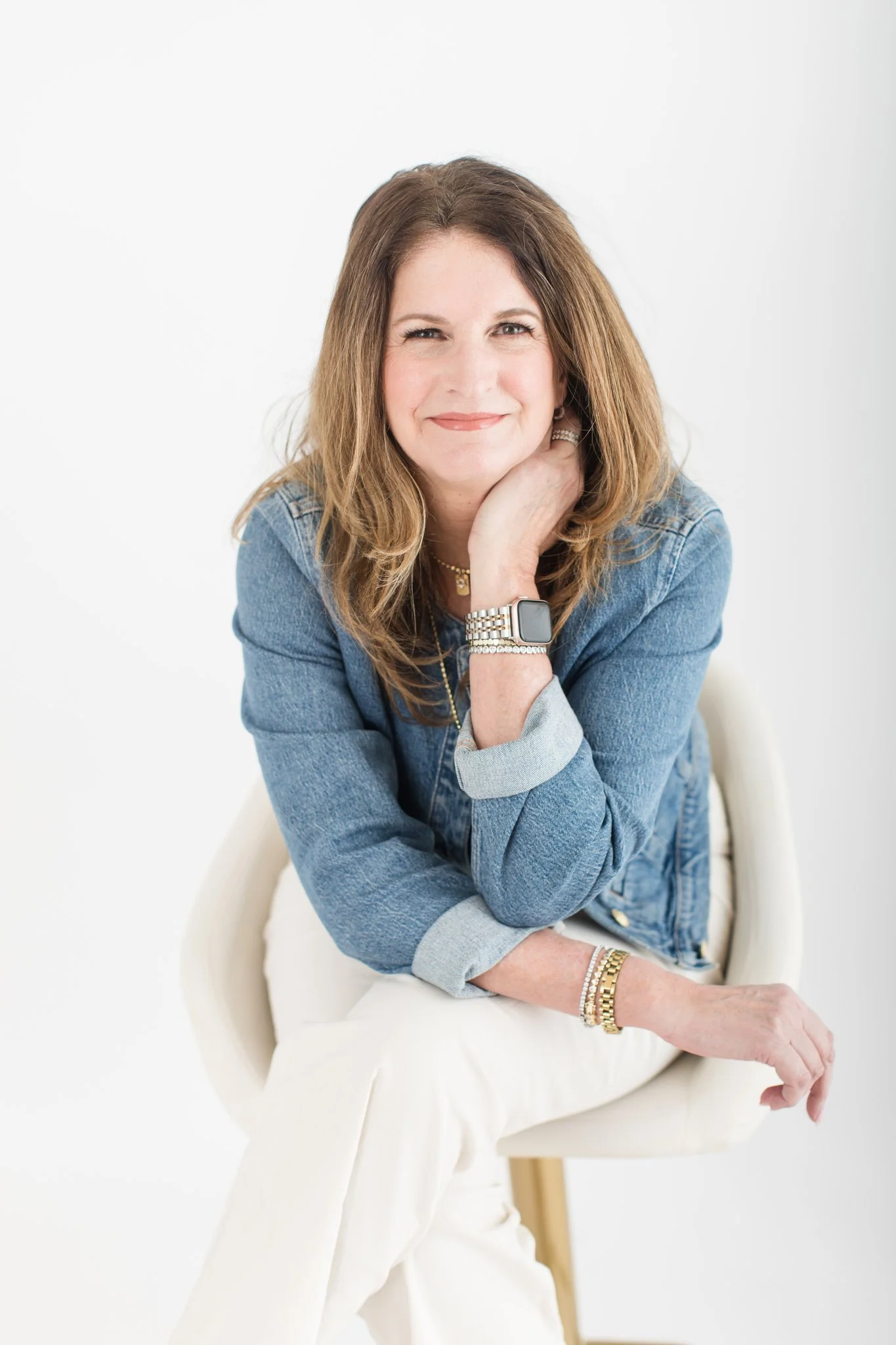 A woman with long, wavy brown hair, wearing a denim jacket and white pants, sitting on a white chair with a white background, smiling and resting her chin on her hand.