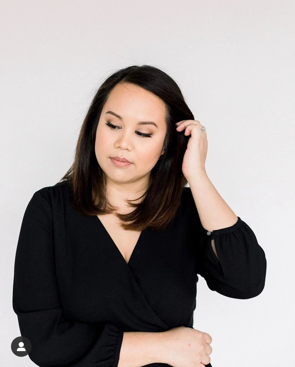 A woman with dark hair wearing a black top, looking down with a thoughtful expression, touching her hair against a plain white background.