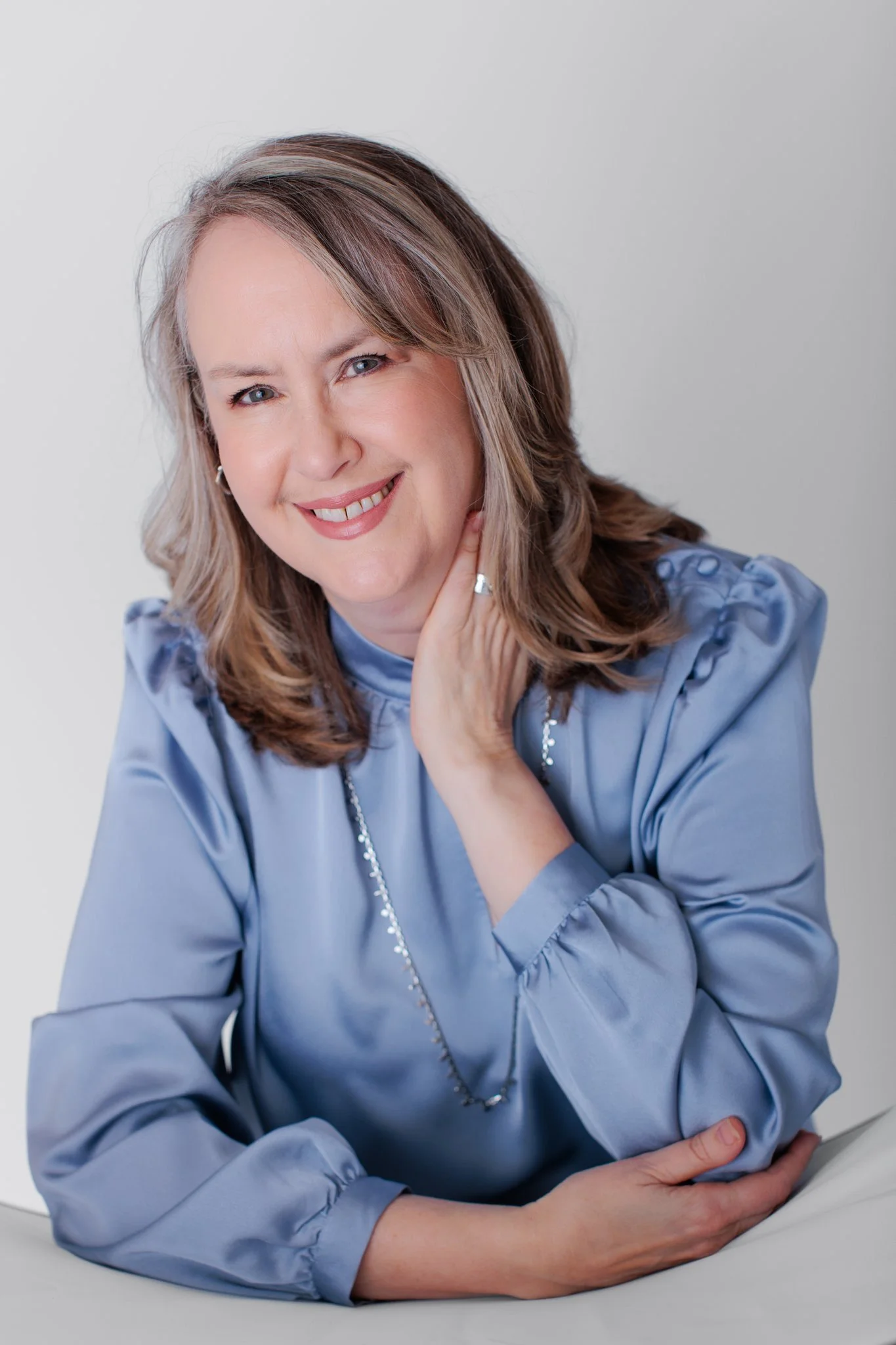 A woman with shoulder-length light brown hair wearing a light blue satin blouse, smiling and touching her neck with one hand, in a professional portrait style against a plain gray background.