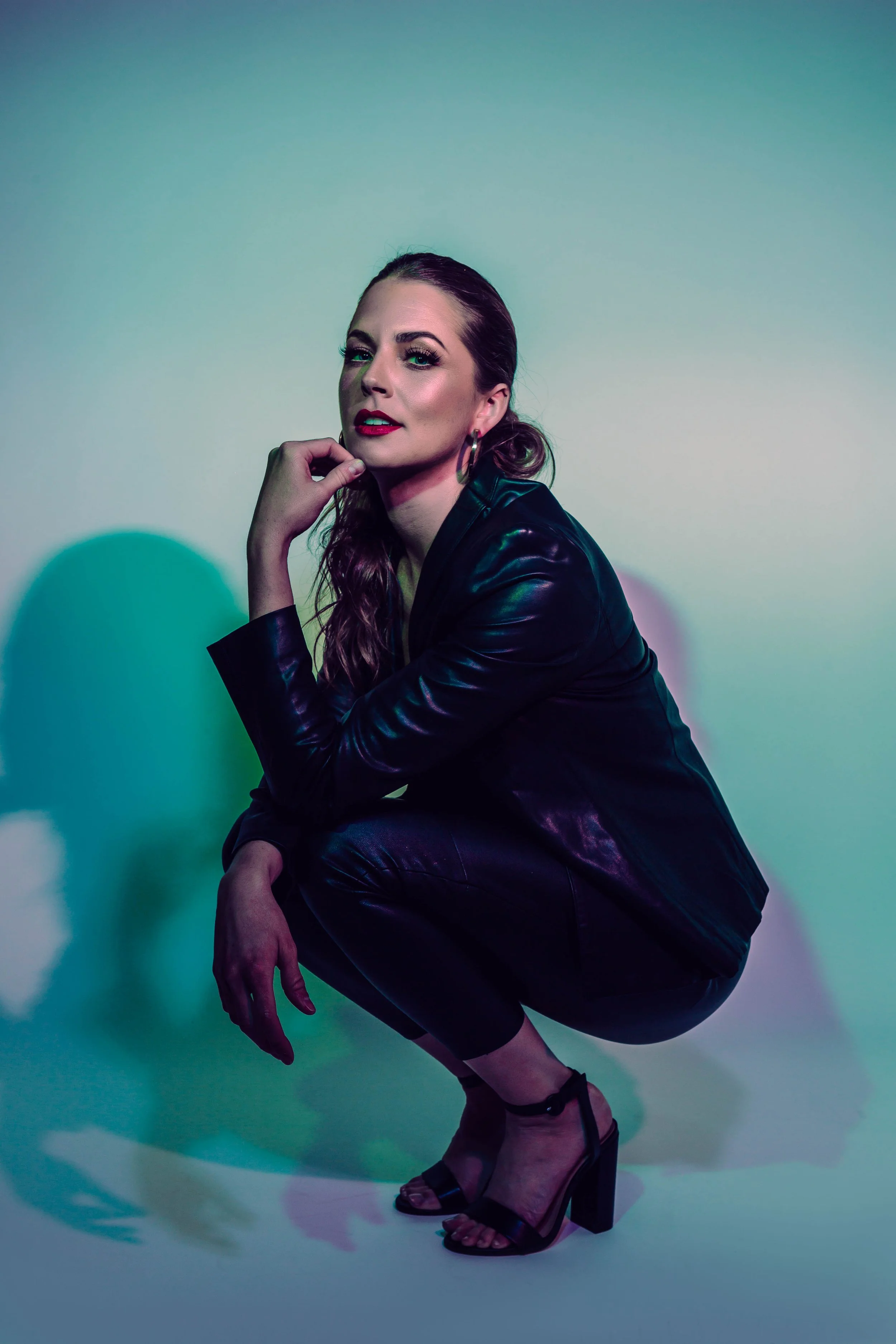 A woman crouching, wearing a black leather jacket and matching pants, with high-heeled sandals, posing against a colorful gradient background.