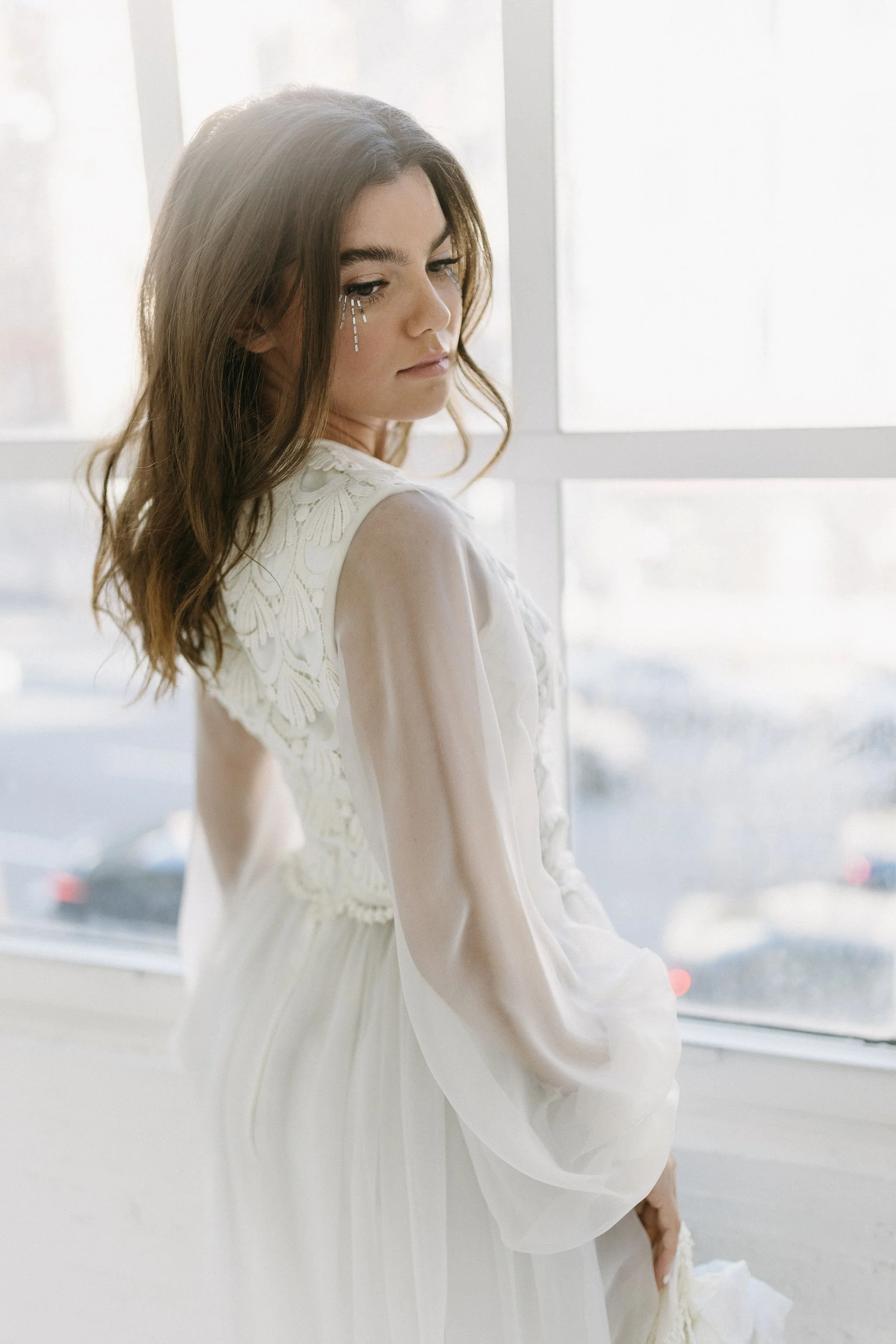 A young woman with wavy brown hair looking down, wearing a white dress with lace details on the shoulders, standing by a large window with city and cars visible outside.