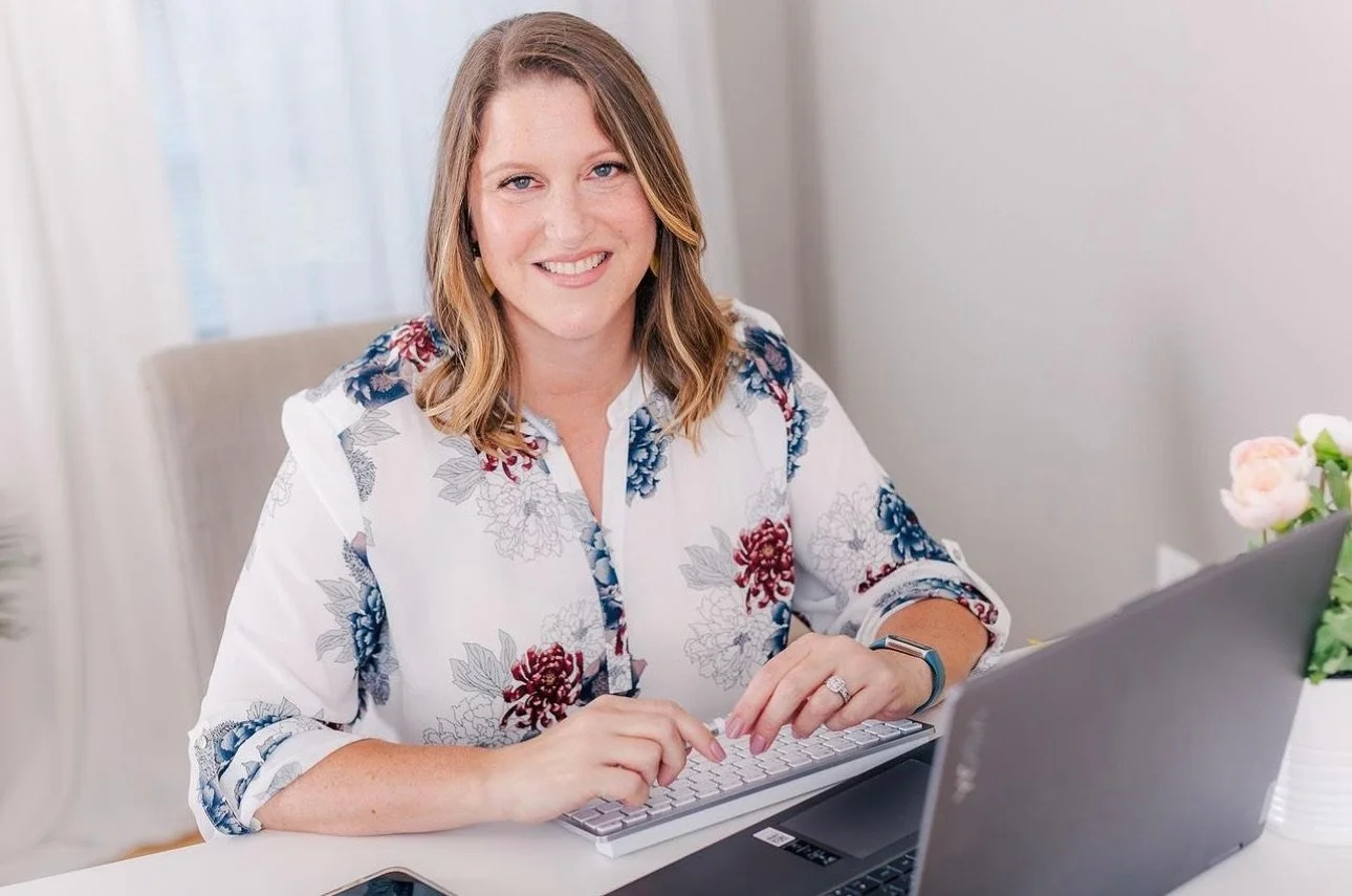 A woman with shoulder-length light brown hair sitting at a desk with a laptop, typing on a keyboard, smiling at the camera, in a bright room with sheer curtains and a small plant on the desk.