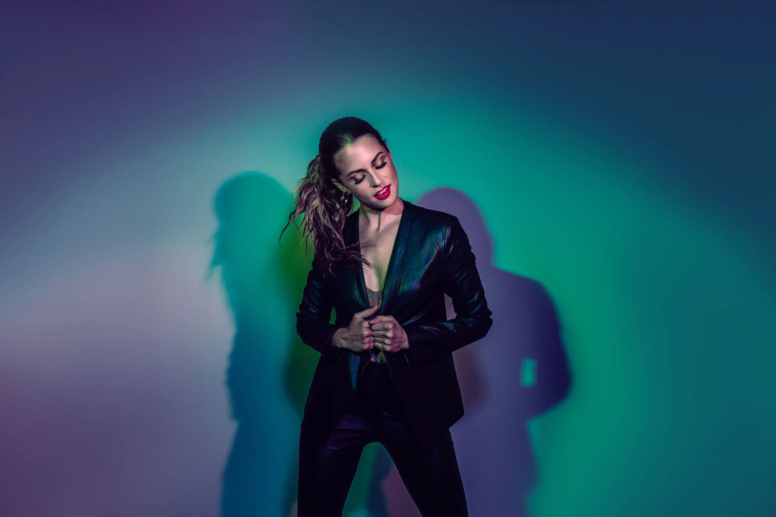 A woman with curly hair wearing a black leather jacket and dark pants poses against a wall with colorful lighting, creating shadow effects.