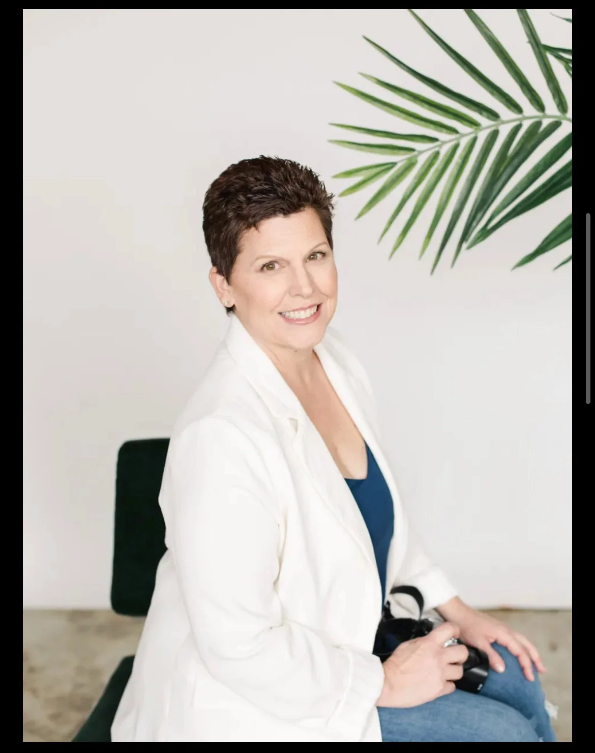 A woman with short, dark hair smiling, wearing a white blazer over a dark top, sitting on a black chair, holding a camera, with green leafy plant in background.