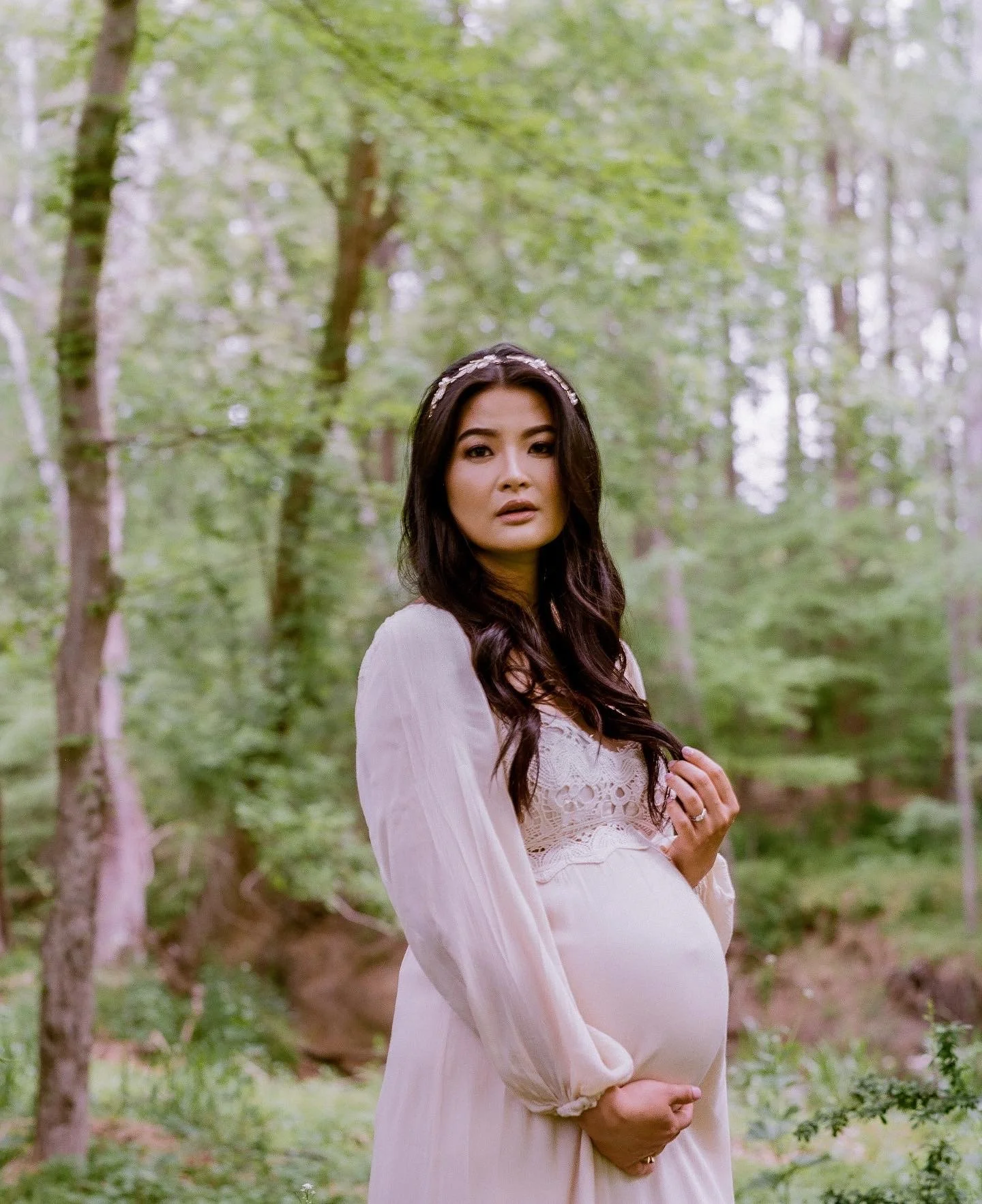 A pregnant woman with long dark hair standing in a forest with green trees, wearing a white dress and a headband, holding her belly, looking at the camera.