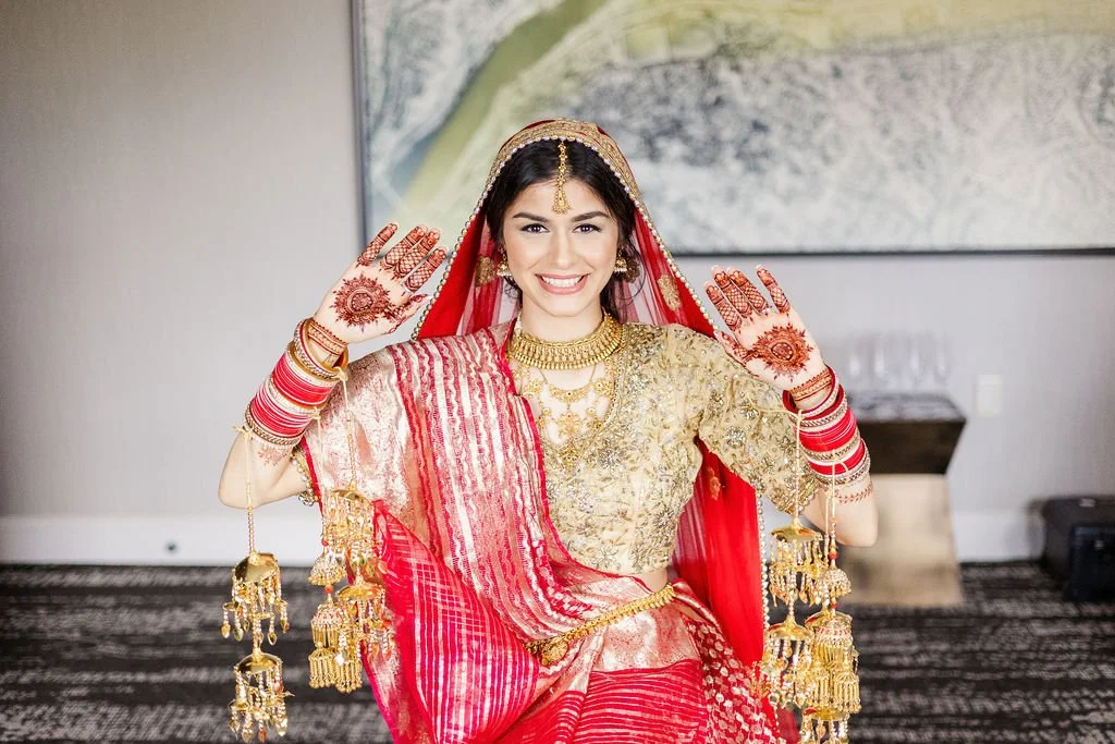 A smiling woman dressed in traditional Indian wedding attire, including a red and gold saree, gold jewelry, and henna on her hands, standing indoors with her hands raised in a wave.