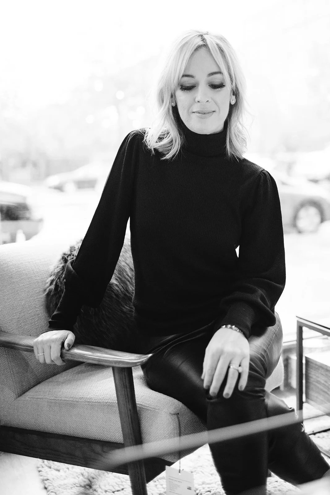 A woman with blonde hair sitting on a modern upholstered armchair inside a cafe or store, wearing a black turtleneck sweater and leather pants, looking down with a subtle smile.