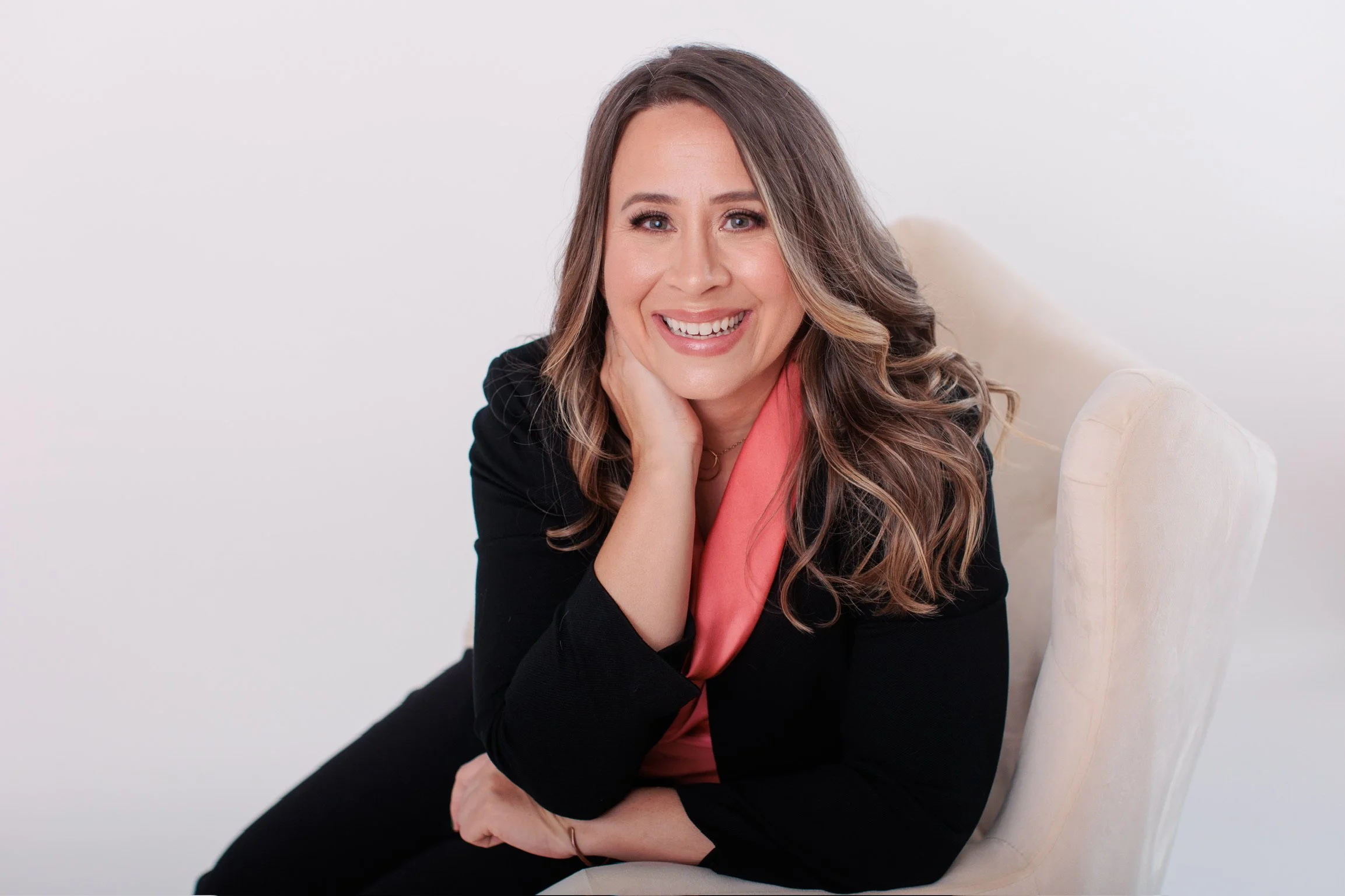 A woman with long wavy brown hair, wearing a black blazer and a pink top, smiling while seated on a beige chair against a plain white background.