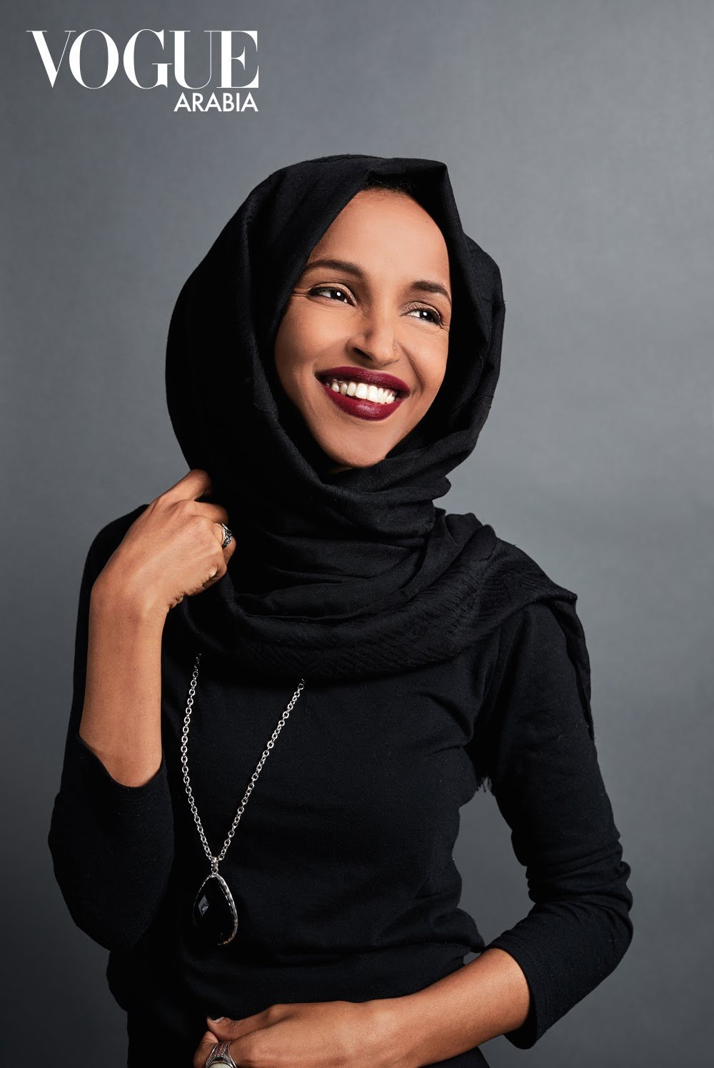 A woman wearing a black headscarf and black top, smiling and posing against a gray background. The Vogue Arabia logo is at the top left corner.