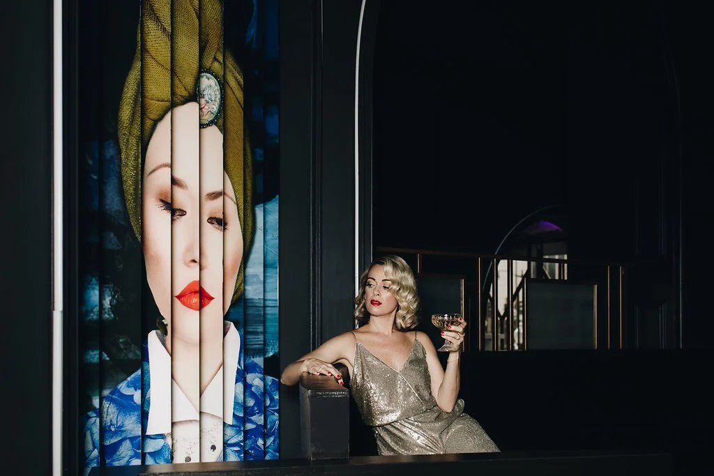 A woman in a gold sequin dress holding a glass of wine, sitting in a dimly lit lounge with a large artistic portrait of a woman with red lips and a green headwrap on the wall behind her.