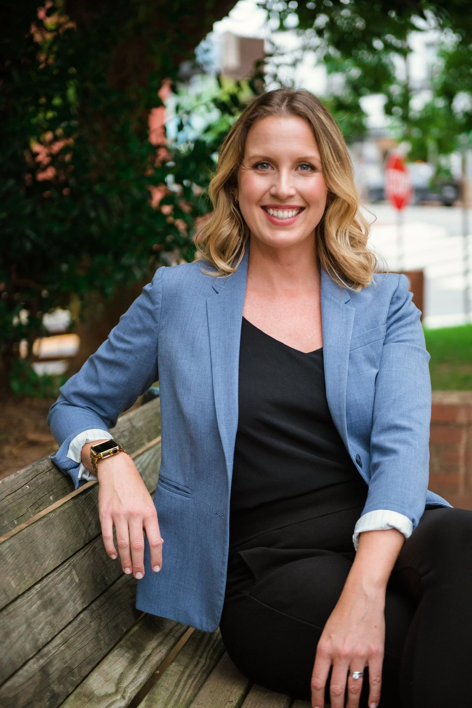 A woman with blonde hair and blue eyes smiling, wearing a blue blazer and black top, sitting on a park bench outdoors with green trees in the background.