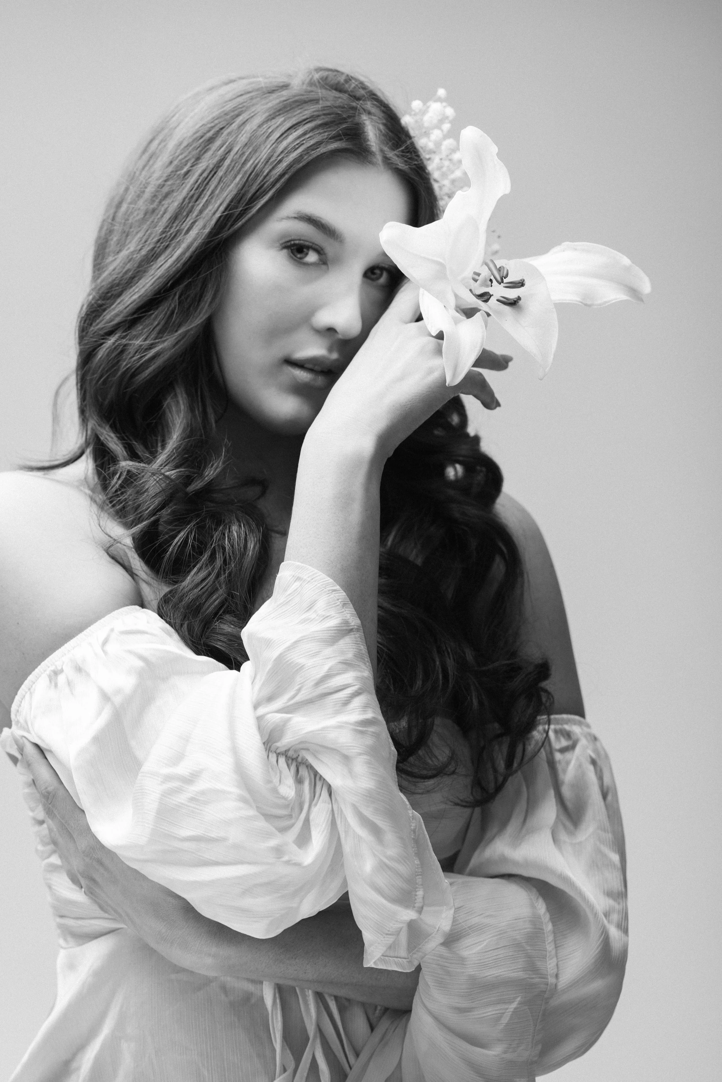 Black and white photo of a woman with long wavy hair, holding a large lily flower near her face, wearing an off-the-shoulder top.