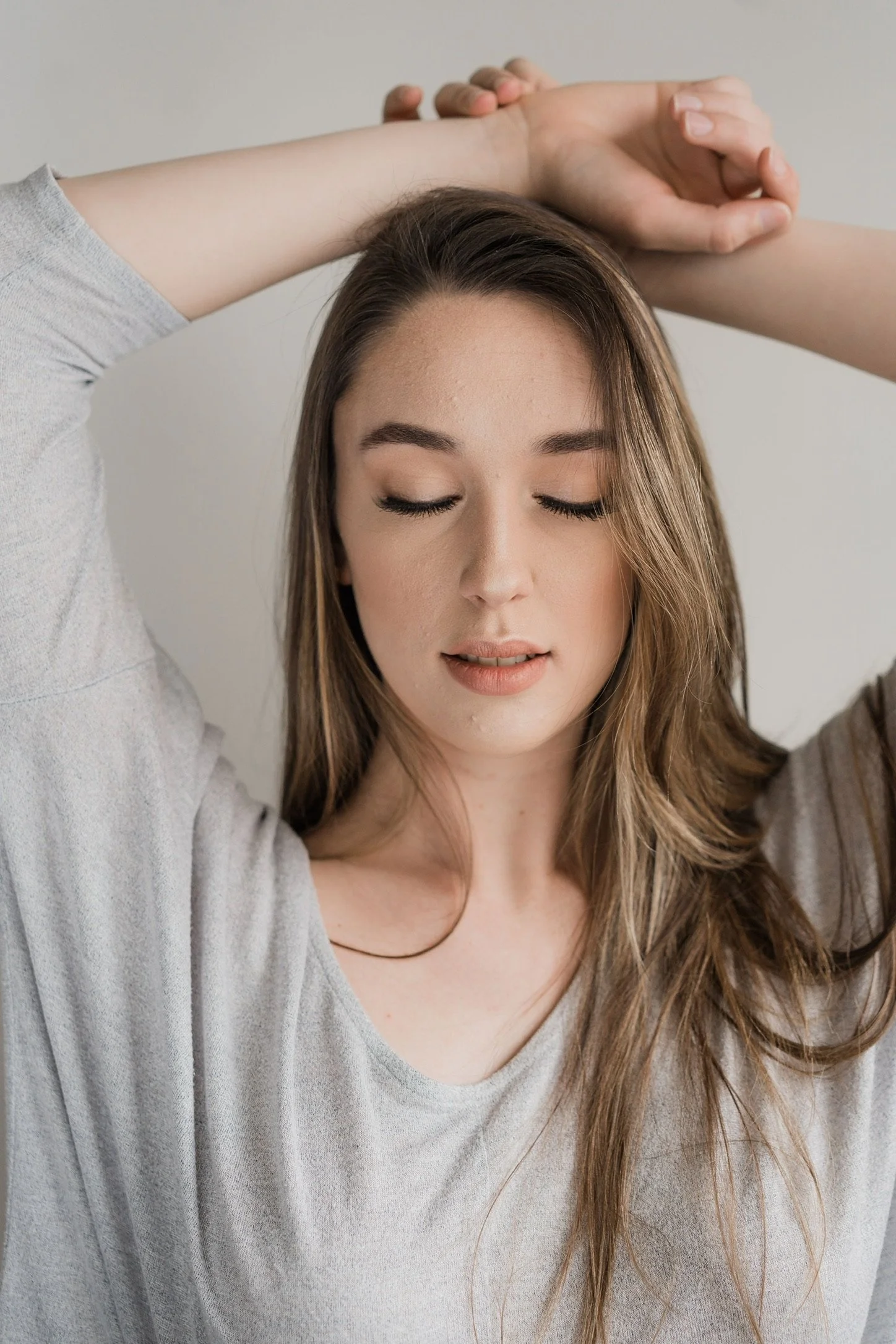 A woman with long, brown hair is posing with her eyes closed and her arms raised above her head, wearing a light gray shirt.