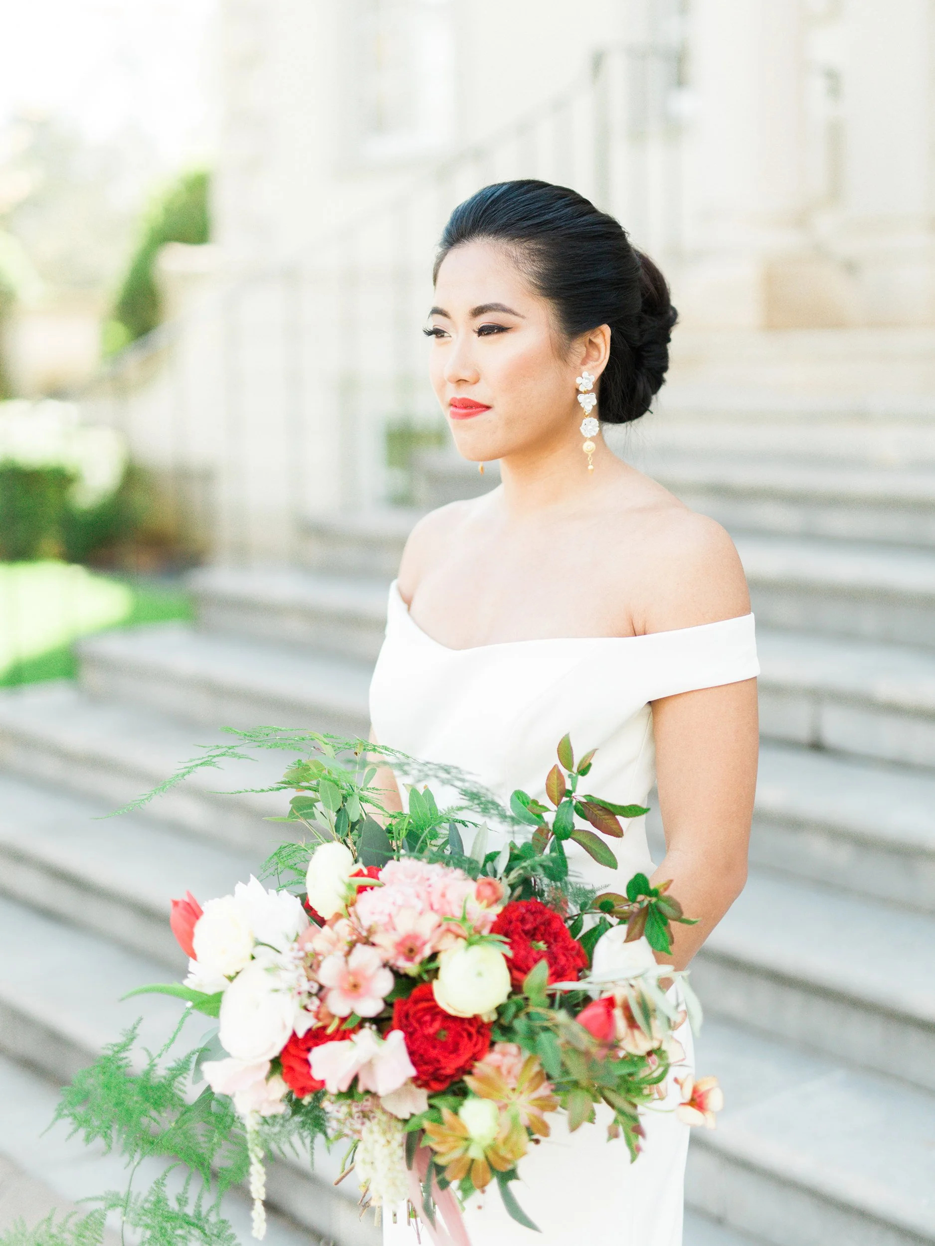 Brunette woman in a white off-the-shoulder dress holding a large bouquet of flowers, standing outdoors in front of steps.