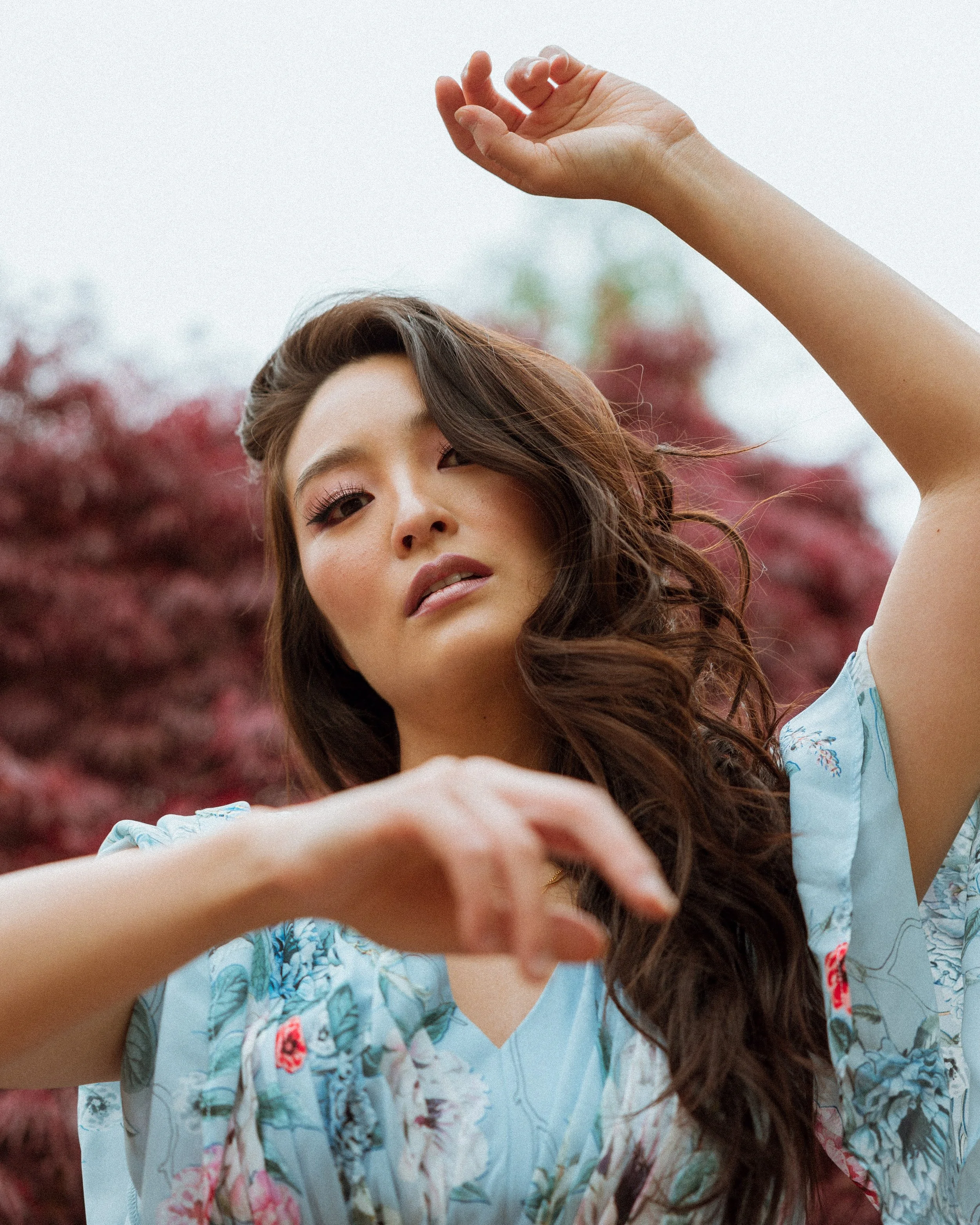 A woman with long, wavy brown hair wearing a blue floral top poses outdoors with blurred trees in the background.