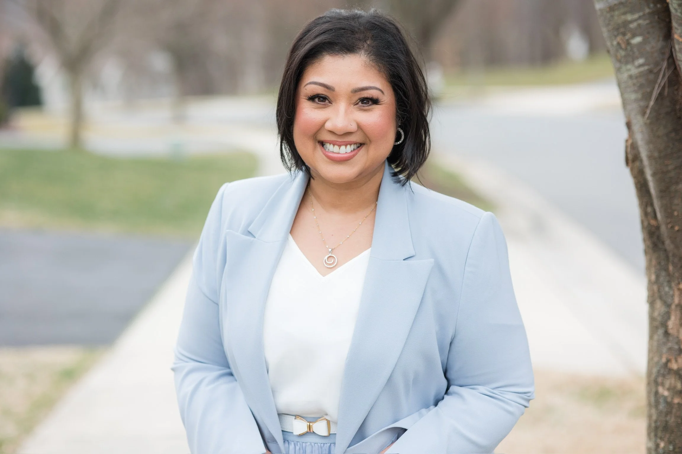 A woman with shoulder-length dark hair smiling outdoors on a sidewalk, wearing a light blue blazer, white top, and gold jewelry, with a tree and a blurred street background.