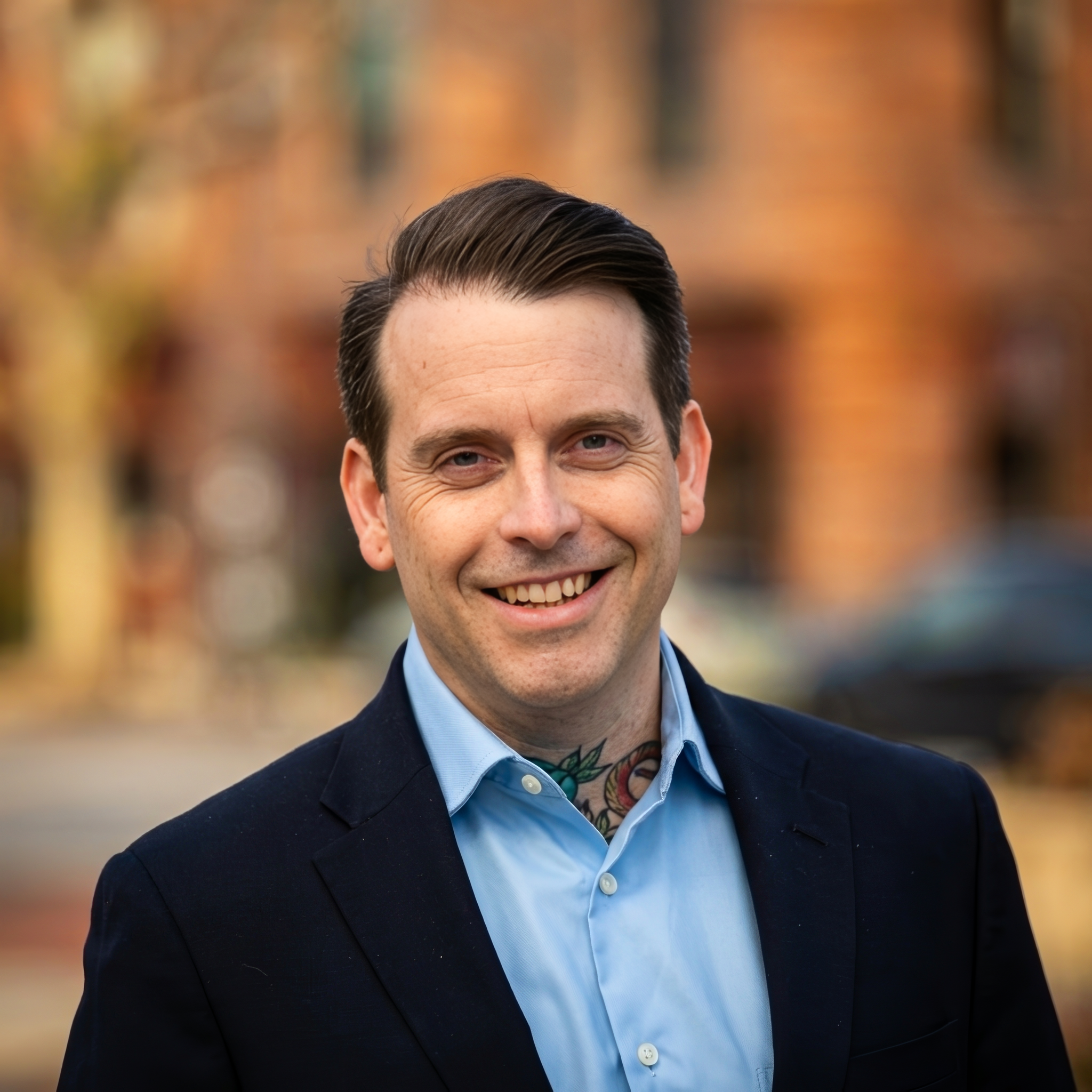 Cameron Moquin with dark hair and tattoos on his neck, wearing a navy blazer and a light blue dress shirt outdoors with blurred trees and buildings in the background.