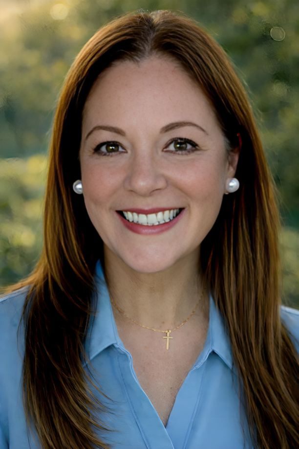 A woman with long red hair smiling outdoors, wearing a light blue collared shirt, pearl earrings, and a gold cross necklace.