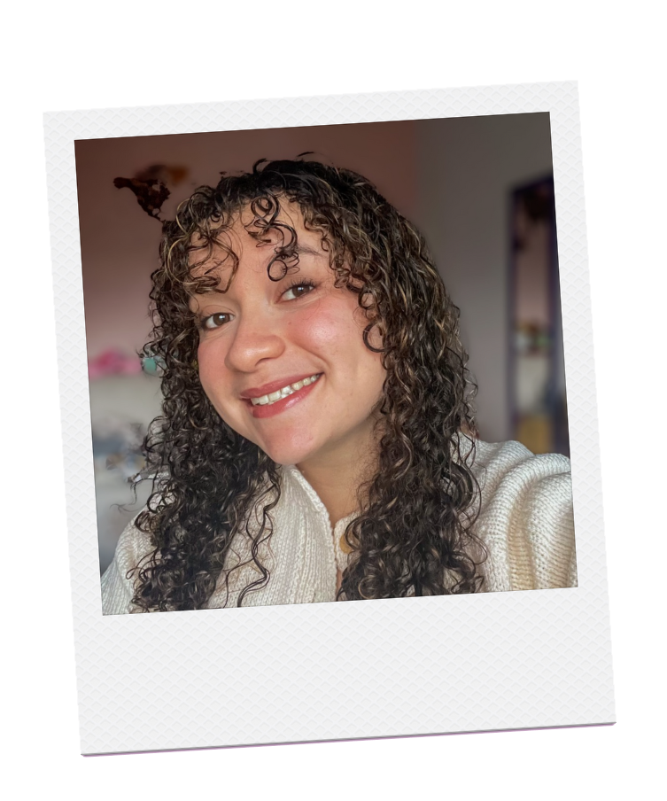 Une femme souriante avec des cheveux bouclés, portant un pull blanc, dans un intérieur domestique.