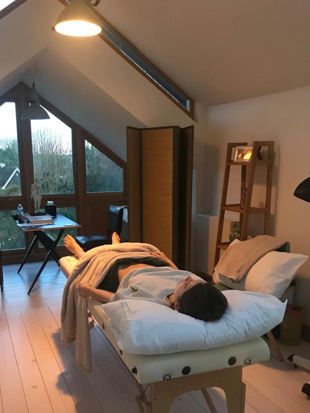 A person lying on a medical examination table in a cozy room with sloped ceiling and large windows. The room has a lamp, wooden furniture, and a view of trees outside.