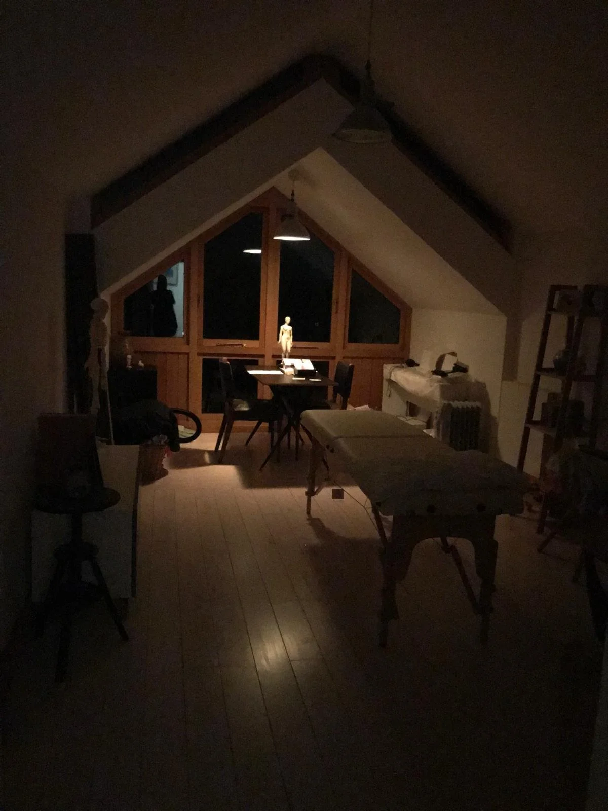 A dimly lit attic room with wooden flooring, a sloped ceiling, and large triangular windows. There is a table with chairs, a mannequin, and various objects including a skeleton model and a lamp.