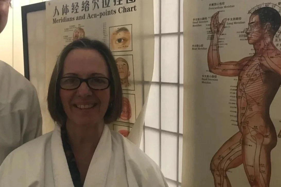 A woman wearing glasses and a white martial arts uniform smiling at the camera. Behind her are posters with human anatomy charts, including acupuncture points and meridians.
