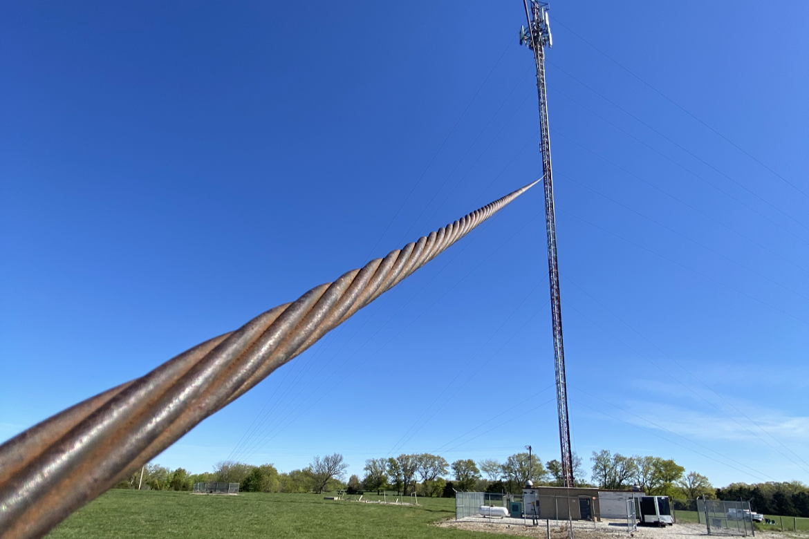 Close-up of a tall, rusted steel pole or cable extending towards a radio tower in the distance under a clear blue sky, with a grassy field and some trees at the horizon.