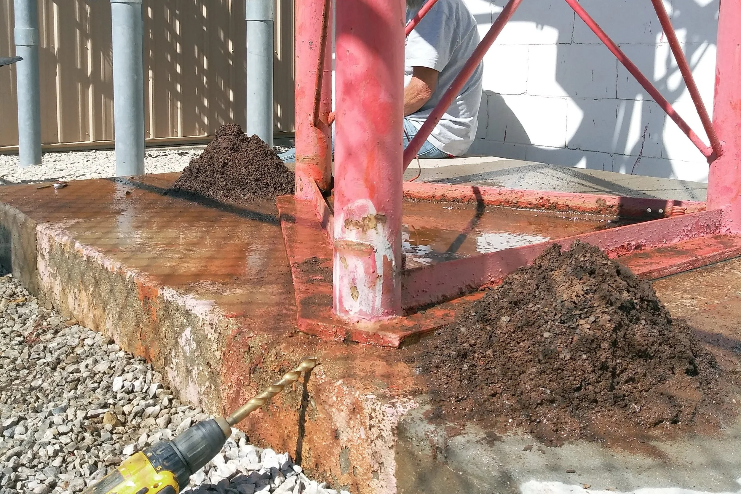 Construction worker at a worksite operating a drill, with dirt piles and scaffolding surrounding a concrete base.