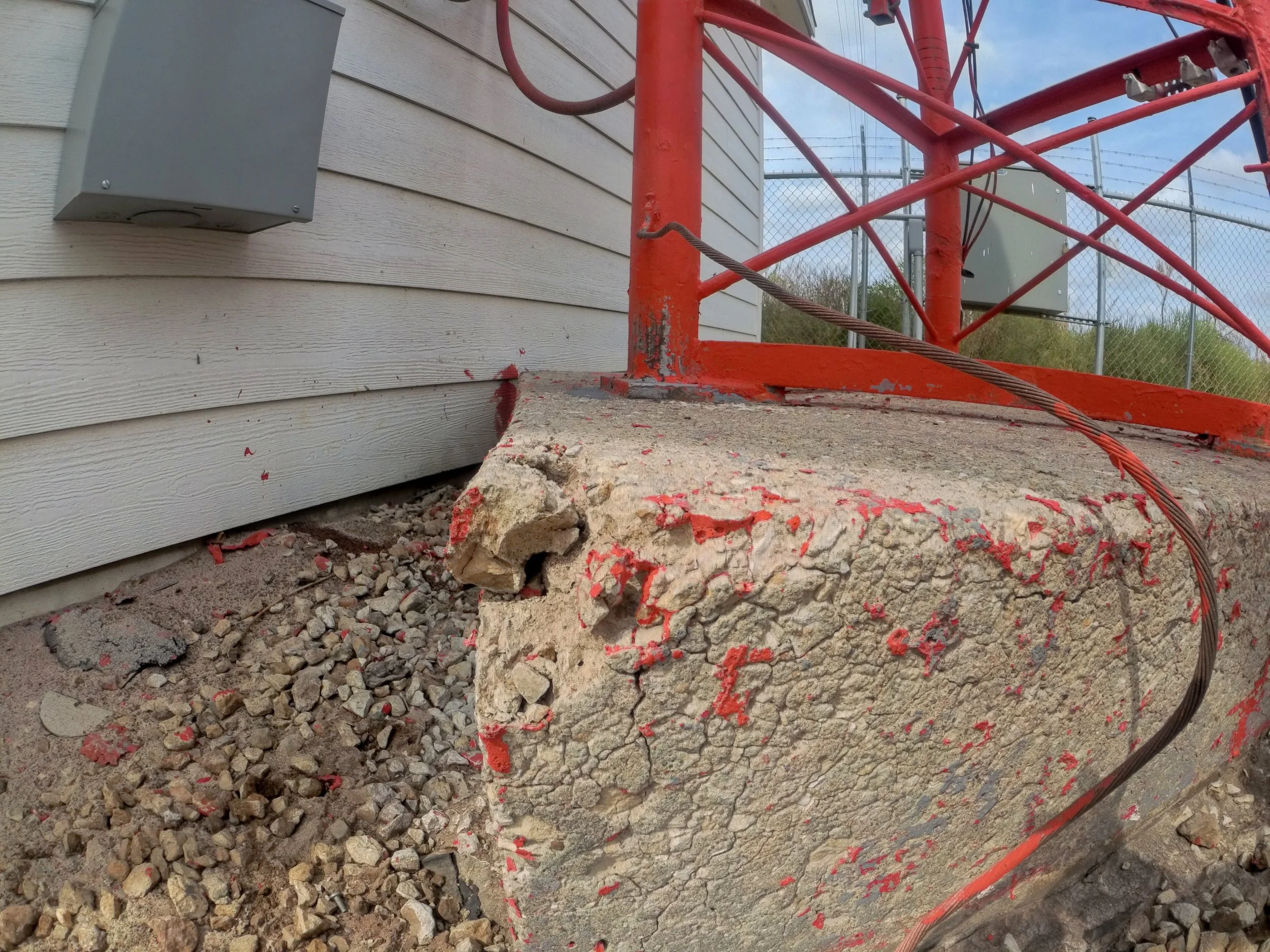 Close-up of a concrete foundation with chipped edges, a chain-link fence in the background, gray electrical boxes on the siding, and red metal scaffolding with a cable running across.