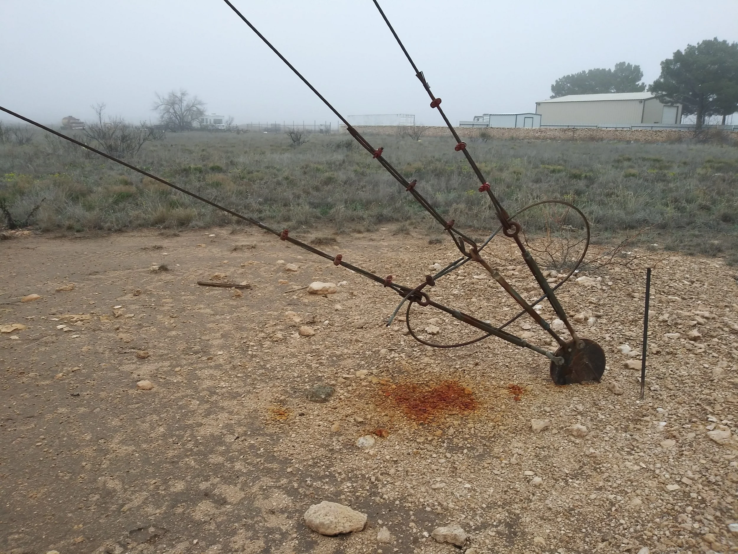 A tilted, rusty metal anchor point on a dirt and gravel ground with some bloodstains nearby, in a foggy outdoor area with sparse vegetation and some buildings and trees in the background.