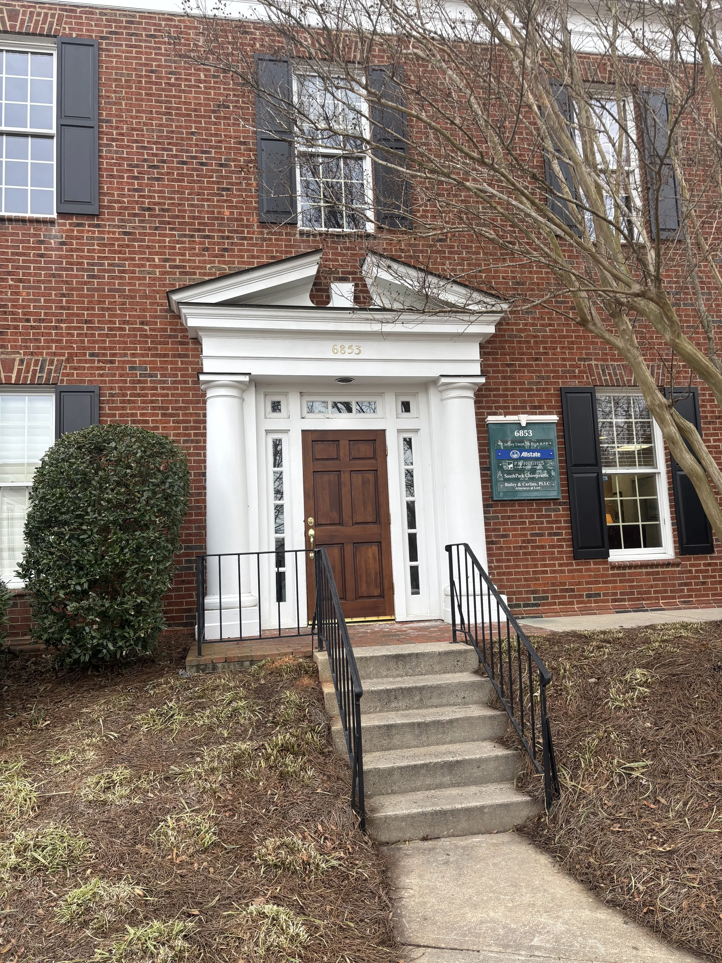 Brick building with white columns framing the front entrance, black shutters on the windows, a brown wooden door, concrete steps with black metal railings leading to the door, a small bush on the left, a signboard on the right, and a leafless tree in front.