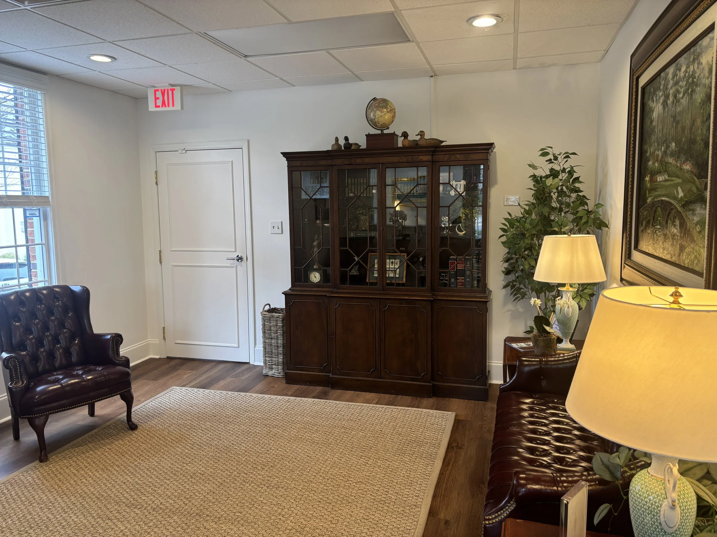 Interior of an office with white walls, a wooden display cabinet, a potted plant, picture frames, a table lamp, and leather armchairs.
