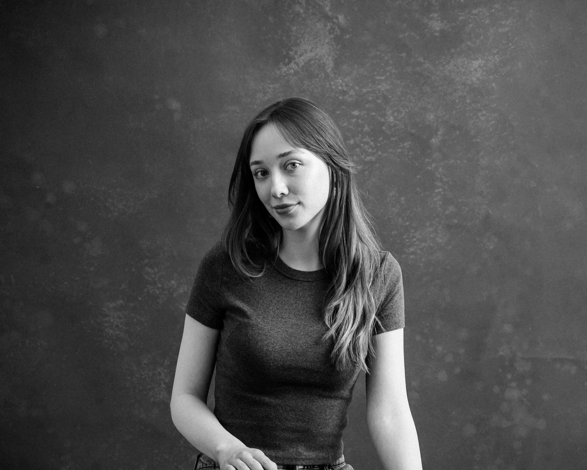 Black and white studio portrait of a young woman with long hair, wearing a short-sleeved shirt, looking at the camera.