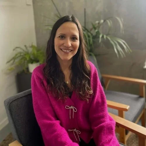 A smiling woman with long, wavy brown hair wearing a bright pink sweater, seated in a modern indoor setting with plants and a wooden chair in the background.