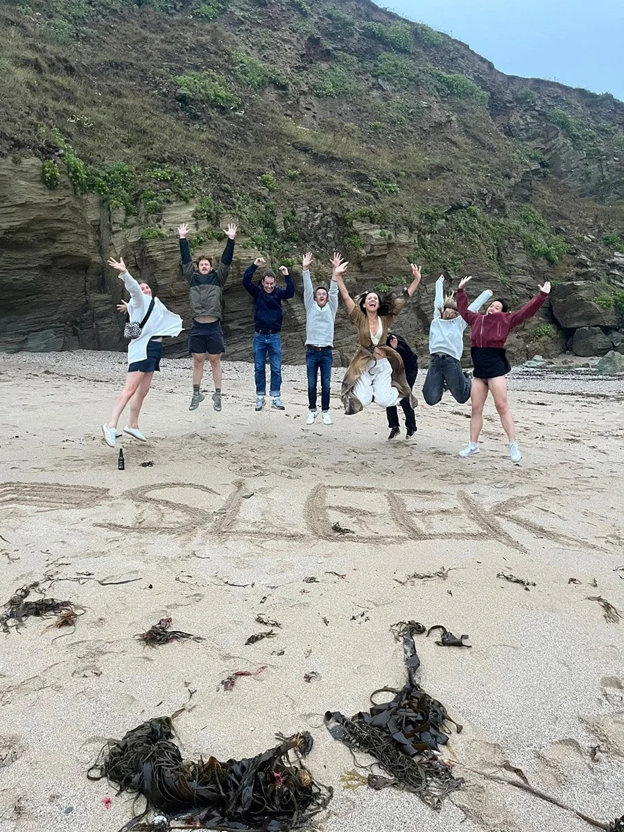 Group of seven people mid-air jumping on a beach with large rocks and cliffs in the background. The word 'GRAD' is written in the sand.