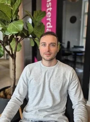 A man with short dark hair and light skin sitting at a table indoors, wearing a light gray long-sleeve shirt, with a green leafy plant behind him.