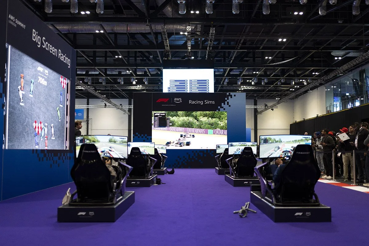 An exhibition area at an AWS event with racing simulators set up for attendees to try virtual racing. There are six racing seats with attached monitors, a large screen displaying a virtual race, and a crowd of people watching on the right side.