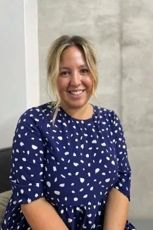A smiling woman with blonde hair wearing a blue dress with white polka dots, standing indoors against a gray concrete wall.